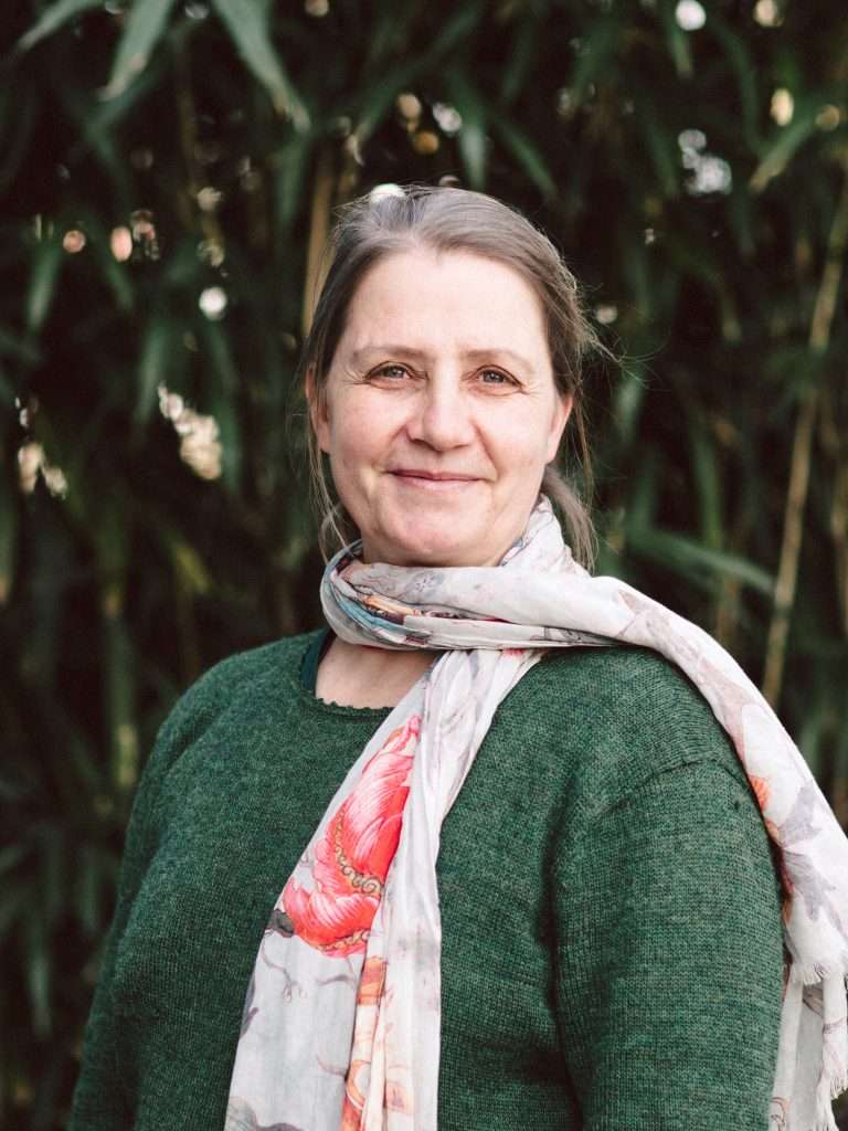 Eine Person steht vor einem Hintergrund aus grünen Blättern. Sie trägt einen dunkelgrünen Pullover und einen hellen Schal mit einem Blumenmuster.